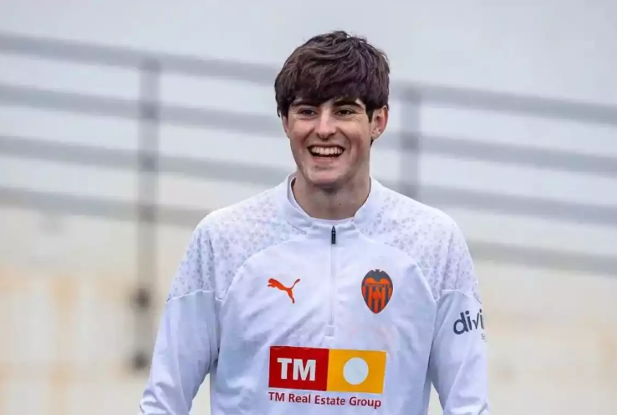A smiling soccer player, Javi Guerra, in a white Valencia CF training jacket stands outdoors.