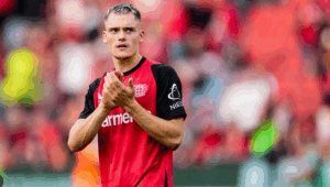 A soccer player in a red and black jersey claps, standing on the field with fans in the background, reminiscent of Bayern Munich celebrations.