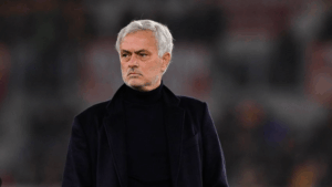 A man with short gray hair in a dark coat stands outdoors, looking serious, evoking Mourinho’s determined presence against a blurred background.