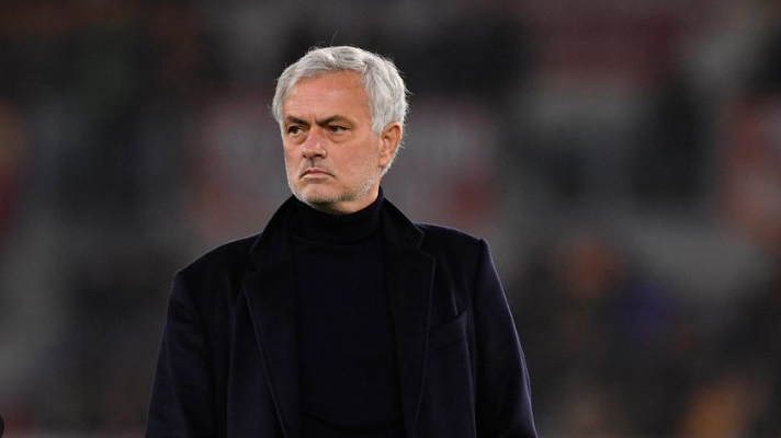 A man with short gray hair in a dark coat stands outdoors, looking serious, evoking Mourinho’s determined presence against a blurred background.