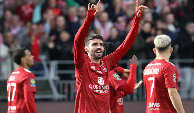 Soccer player in red jersey celebrates with raised hands after a Rennes victory; teammates and crowd visible in background.