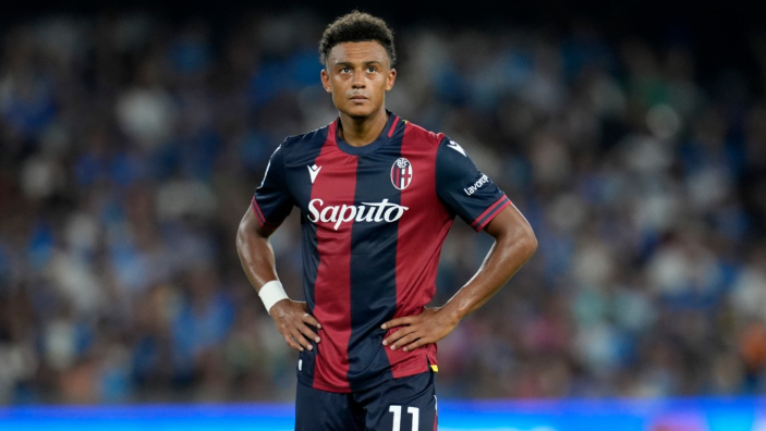 Ndoye, a soccer player in a red and blue Bologna jersey, stands on the field with hands on hips during a match against Napoli.