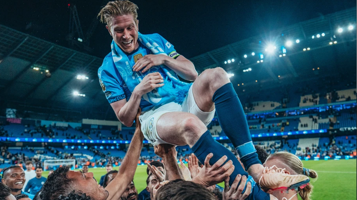 Soccer player lifted joyfully by teammates on the field, celebrating a farewell in the stadium at night—a scene reminiscent of De Bruyne's memorable moments with Manchester City.
