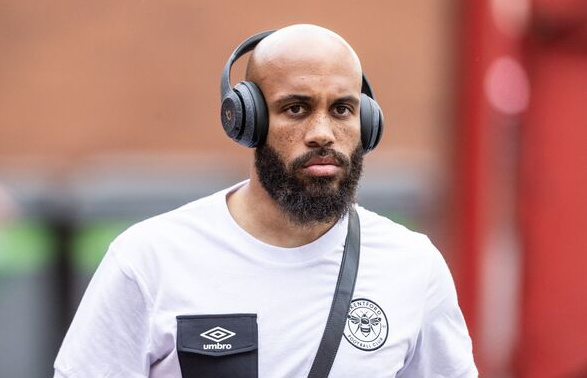 A man with a beard wearing headphones and a white t-shirt walks outdoors, sporting a Brentford cap and showing his support for Mbeumo.