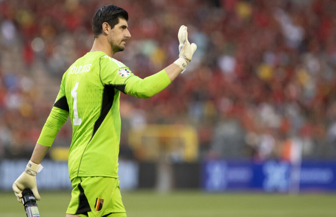 Goalkeeper in a green kit, reminiscent of Real Madrid’s Courtois, waving during a soccer match, with a blurred crowd in the background.