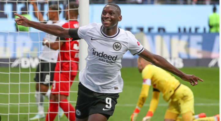 Kolo Muani, in a white Eintracht Frankfurt jersey, celebrates a goal with arms outstretched near the goalpost amid transfer rumors linking him to Man United.
