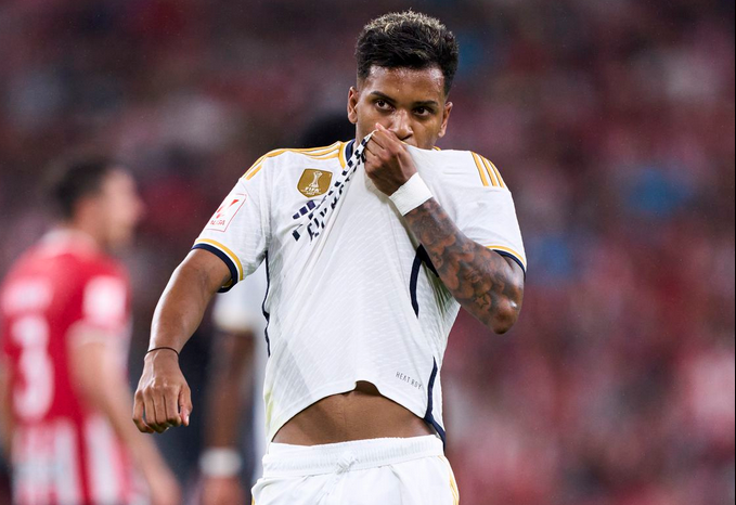 Soccer player Rodrygo kisses his team’s badge on his jersey during a match, with blurred opponents in the background, celebrating a proud Real Madrid moment.
