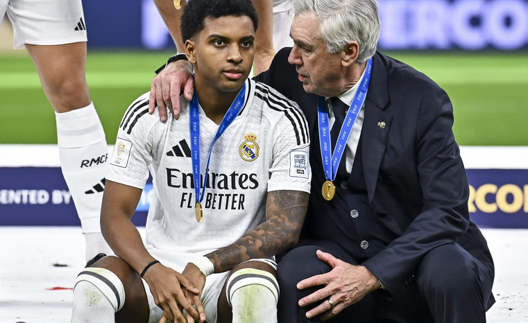 Soccer player Rodrygo and coach in Real Madrid uniforms sit together wearing gold medals after a match.