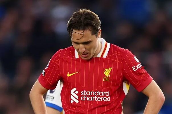 A football player in a red Liverpool jersey, reminiscent of Serie A star Chiesa, looks down with hands on hips during a match.