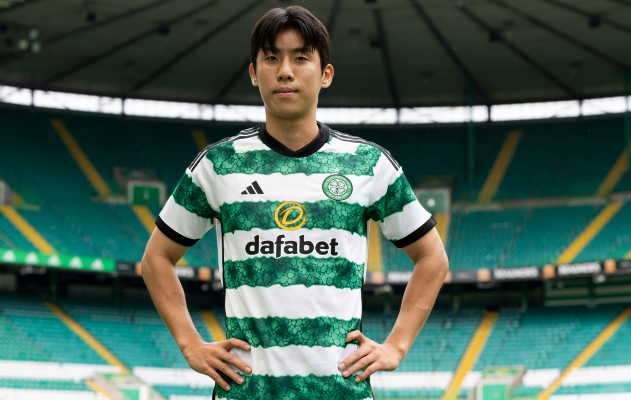 A South Korean midfielder in a green-and-white Celtic FC jersey stands with hands on hips in an empty stadium.