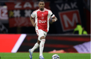 Ajax football player Hato in red and white kit, known as a Dream Defender, running with the ball on the soccer field during a match.