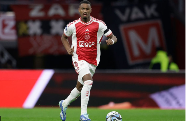 Ajax football player Hato in red and white kit, known as a Dream Defender, running with the ball on the soccer field during a match.