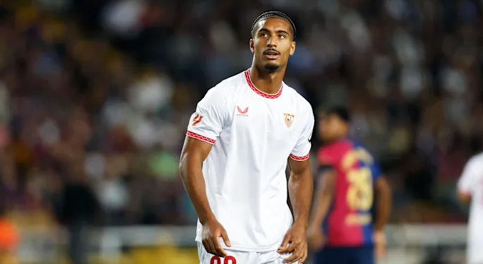 French defender Badé, wearing a white Sevilla FC uniform, stands on the field during a match, looking to the side.