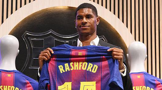 A man in a suit holds a Barcelona jersey with "Rashford 10" printed on the back, hinting at a possible Barcelona loan move for Rashford to showcase his skills at Camp Nou.