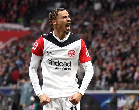 A soccer player in a Bayern Munich jersey, possibly Lee Hyun-ju, juggles a ball on a field near empty stadium seats.