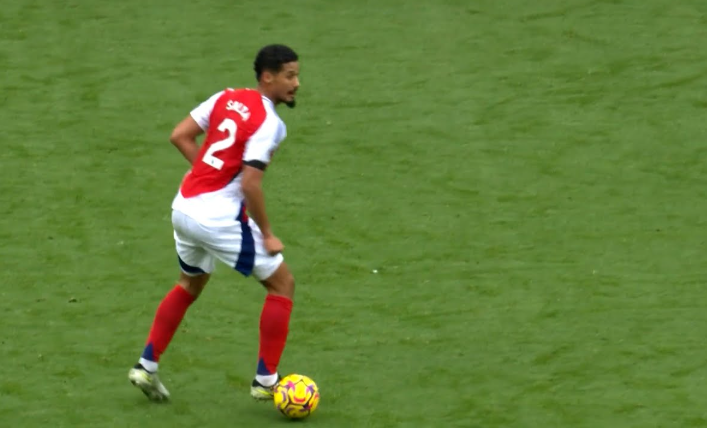 Soccer player in a red and white jersey dribbles the ball on a green field, closely watched by Konate.