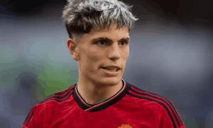 A male soccer player, resembling Garnacho, stands on the Manchester United training ground in a red jersey, looking to the side with short hair featuring blond highlights.