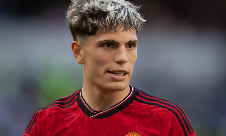 A male soccer player, resembling Garnacho, stands on the Manchester United training ground in a red jersey, looking to the side with short hair featuring blond highlights.