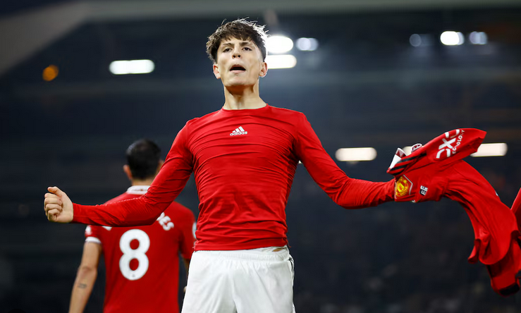 Manchester United’s Garnacho, in a red jersey, celebrates by raising his arms and holding his shirt, with another player visible in the background on the field.