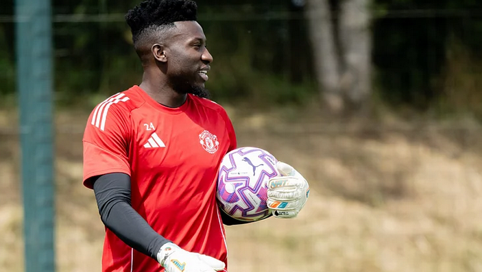 A smiling Man United goalkeeper, Onana, in a red jersey holds a purple and white ball during pre-season outdoor training.