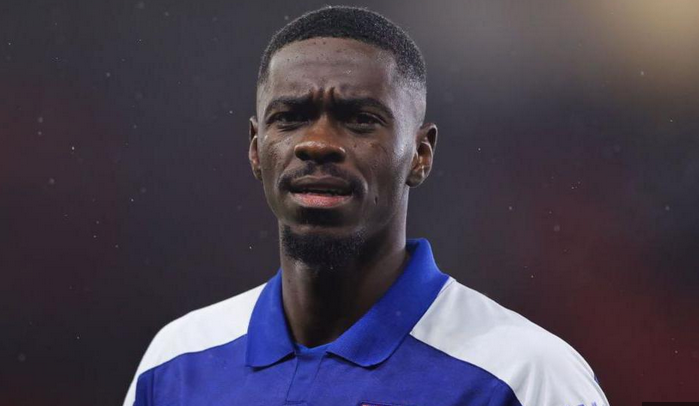 A male soccer player in a blue and white jersey stands on the field, looking forward with a serious expression, reminiscent of Axel Tuanzebe during his days at Man Utd.
