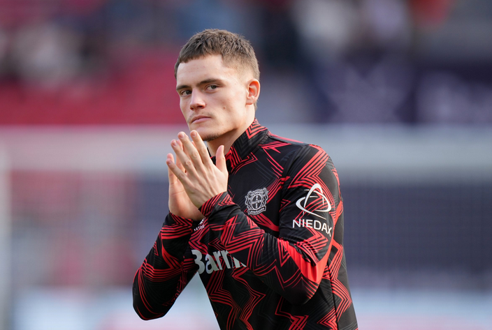 Soccer player in a black and red jacket stands with hands together, looking focused before a match—reminiscent of Wirtz before Liverpool vs Man Utd clashes.