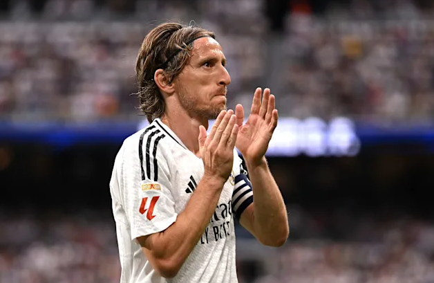 Soccer player in a white Real Madrid jersey claps with both hands on the field, crowd blurred in the background—a scene reminiscent of Modric’s elegance in Italian Football.
