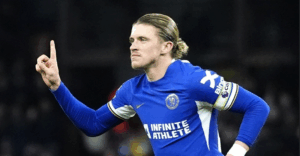 Gallagher, in a blue jersey, celebrates with a determined expression, raising one finger after scoring in a thrilling Premier League match.