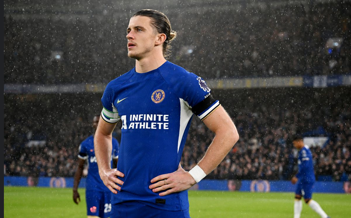 A soccer player in a blue Chelsea jersey stands with hands on hips during a rainy Premier League match.
