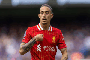 Liverpool defender Tsimikas is on the field in a red jersey, touching the club badge on his chest.