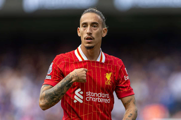 Liverpool defender Tsimikas is on the field in a red jersey, touching the club badge on his chest.