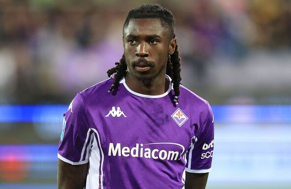A soccer player in a purple Fiorentina jersey stands on the field, looking to the side amid swirling Deadline Drama around a potential Kean transfer to Man United.