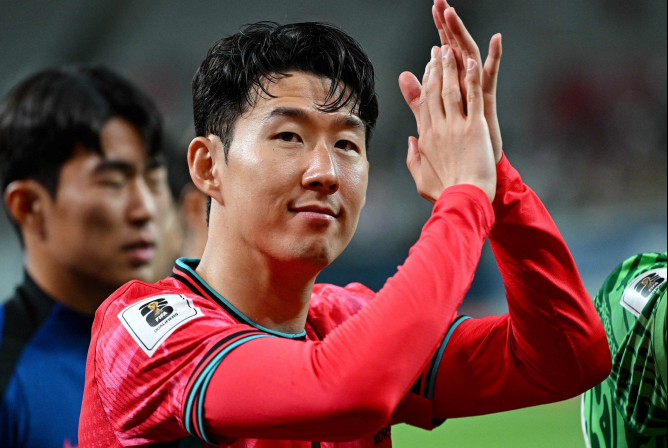 A soccer player in a red jersey claps while looking at the camera, reminiscent of Son Heung-Min during Tottenham’s Golden Era, with teammates in the background.