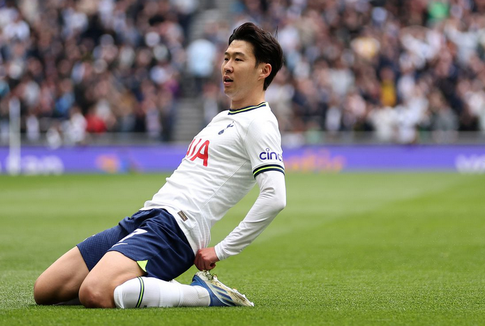 Son Heung-Min, in a white Tottenham jersey, slides on his knees celebrating a goal on a green field as the crowd cheers—capturing a moment from the club’s Golden Era.