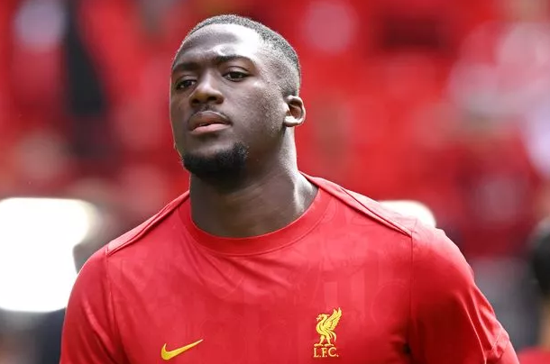 Footballer Konate in a red Liverpool FC shirt with a gold crest, standing on a field with a blurred red background.