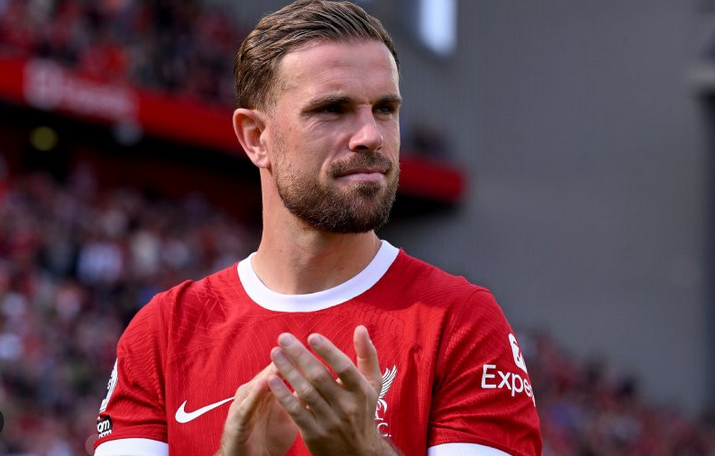 A male football player in a red Liverpool jersey, possibly Henderson, claps hands on the field after a Champions League match, with a stadium in the background.