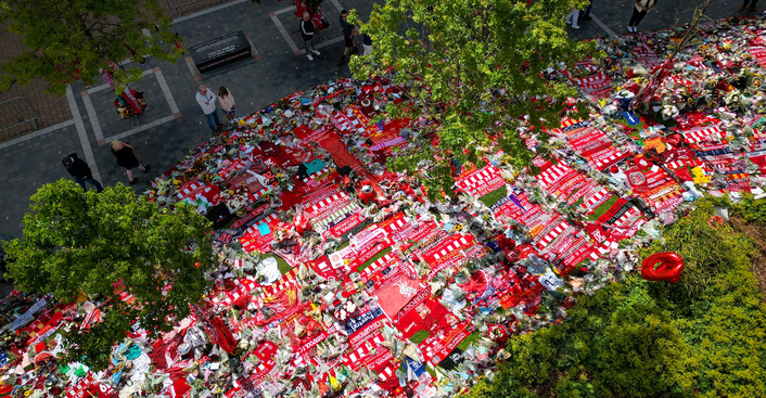 Aerial view of red scarves and tributes laid out under trees near Anfield, with people standing nearby in a parking area.