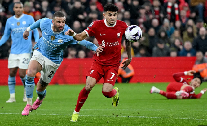 Liverpool’s Diaz and a Bayern player chase the ball during a match, with teammates and the crowd in the background.