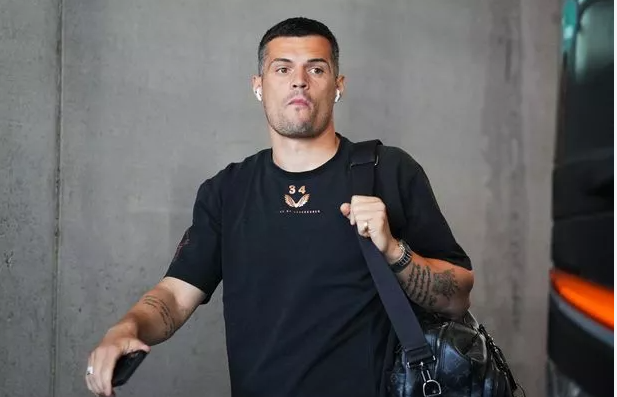 Man in a black t-shirt with tattoos carries a black bag and holds a phone, standing by a concrete wall—his look as sharp as Premier League style on match day.