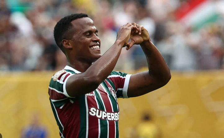 Soccer player in a striped jersey makes a heart gesture with his hands and smiles on the field, celebrating like Wolves star Jhon Arias in a Premier League match.