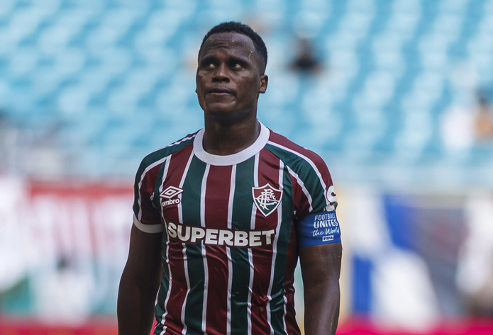 Soccer player Jhon Arias in a maroon, green, and white striped jersey on the field, with a blurred stadium background, reminiscent of Premier League action.