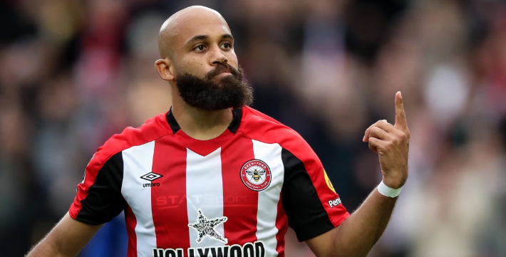 Brentford's Mbeumo, in a red and white jersey, raises his right hand with a look of determination on the field amid transfer rumors linking him to Manchester United.