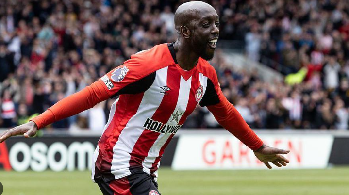 Brentford striker in red and white jersey celebrates with arms outstretched on the field in front of a cheering crowd.