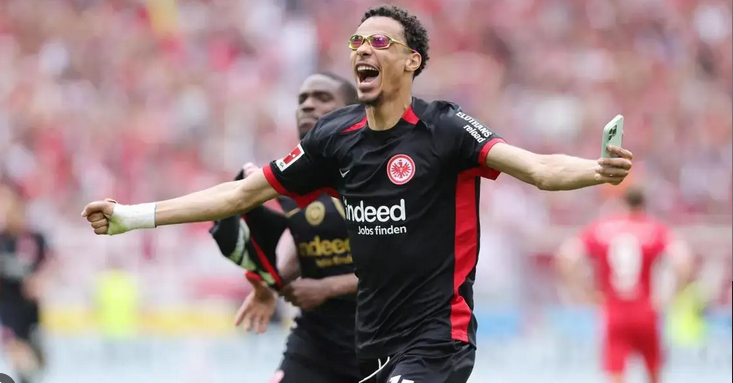 Frankfurt striker Ekitike, in a black Eintracht Frankfurt kit, celebrates with arms outstretched and holds a phone on the field.